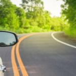 A close-up of a car’s side-view mirror reflecting greenery while driving on a winding road surrounded by lush trees and vegetation.