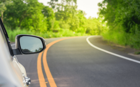 A close-up of a car’s side-view mirror reflecting greenery while driving on a winding road surrounded by lush trees and vegetation.